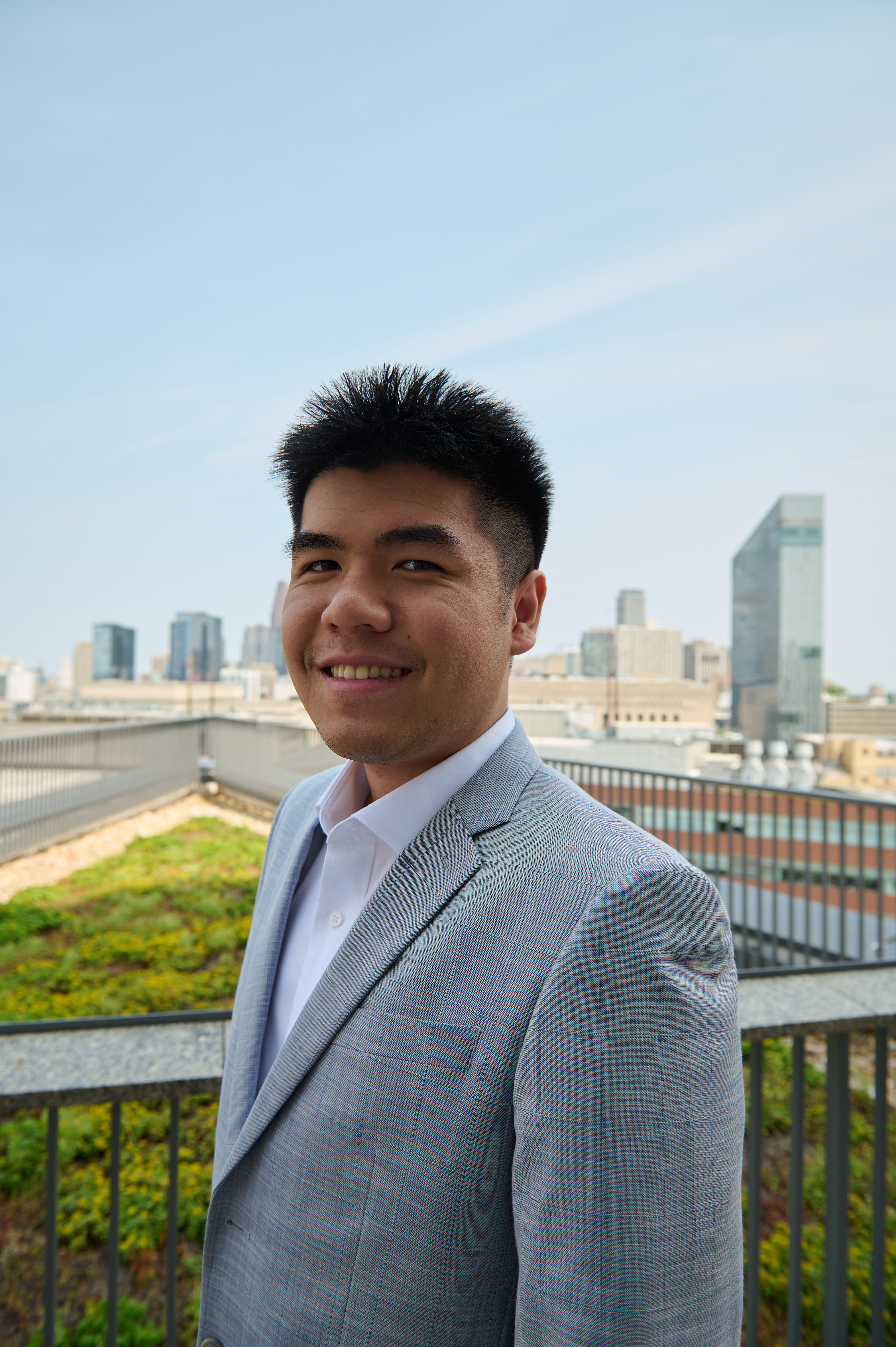 A young man stands outside in a grey suit with the Philadelphia skyline in the background.